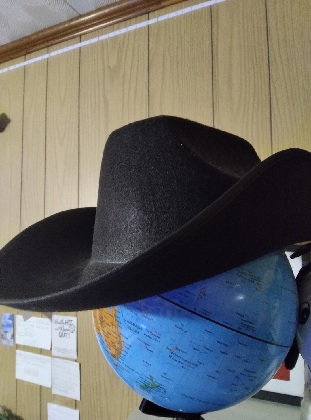 A black cowboy hat rests on a blue globe. In the background, a wooden wall has notes pinned to it, one of which reads 'You'll HIT it when you QUIT!'. The globe shows geographical names like Melanesia, Coral Sea, Fiji, Polynesia, and South Pacific.