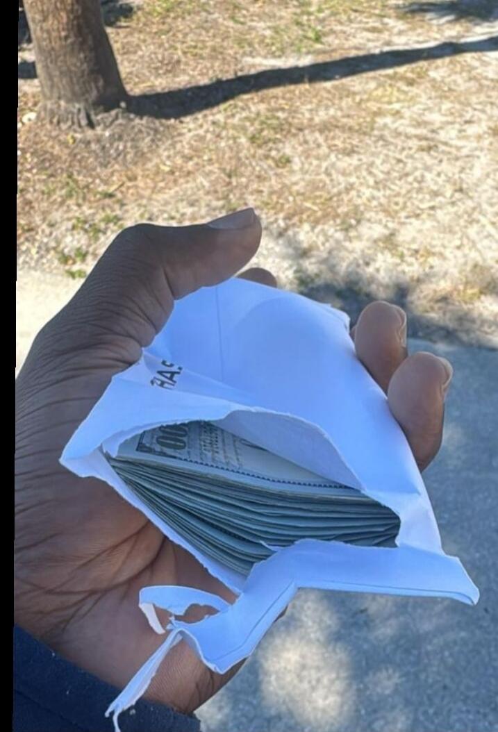 A hand holding an envelope with a stack of US dollar bills, specifically $100 bills. Text visible on the envelope and bills includes 'HAS' and '100'.