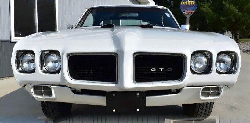 White classic car (Pontiac GTO) front view with distinctive grille and dual headlights. The GTO badge is visible on the grille.