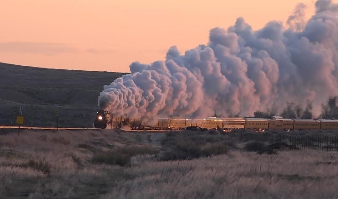 A steam locomotive with a long train emitting large smoke plumes across a barren landscape at sunset.