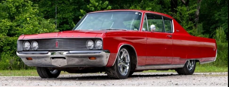 A vibrant red classic American car, possibly a Chrysler Imperial or similar model from the late 1960s, is parked on a gravel road. The car features a prominent chrome grille, dual headlights, and shiny chrome wheels. Lush green foliage forms the background.