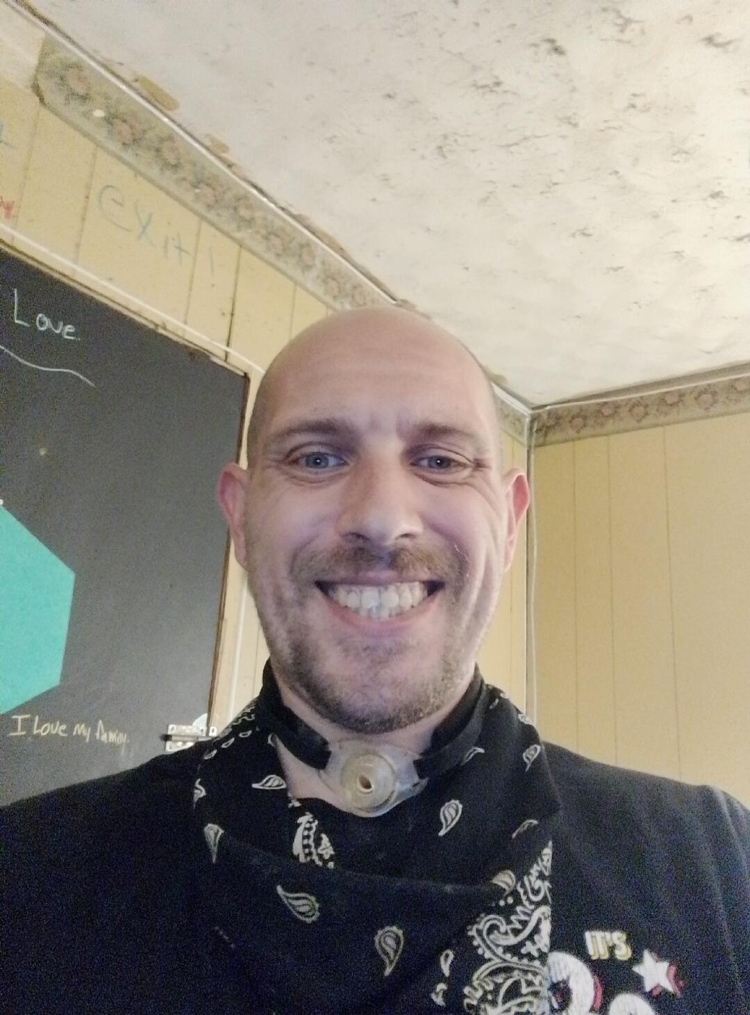 A man with a medical device around his neck smiles broadly at the camera. Text on the wall behind him reads 'Love', 'I Love my family.', and 'Exit!'.