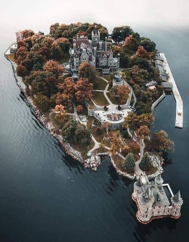 An aerial view of a castle-like building on a forested island, surrounded by water. The island features winding paths, a central courtyard, and a statue on a rocky outcrop near the water.