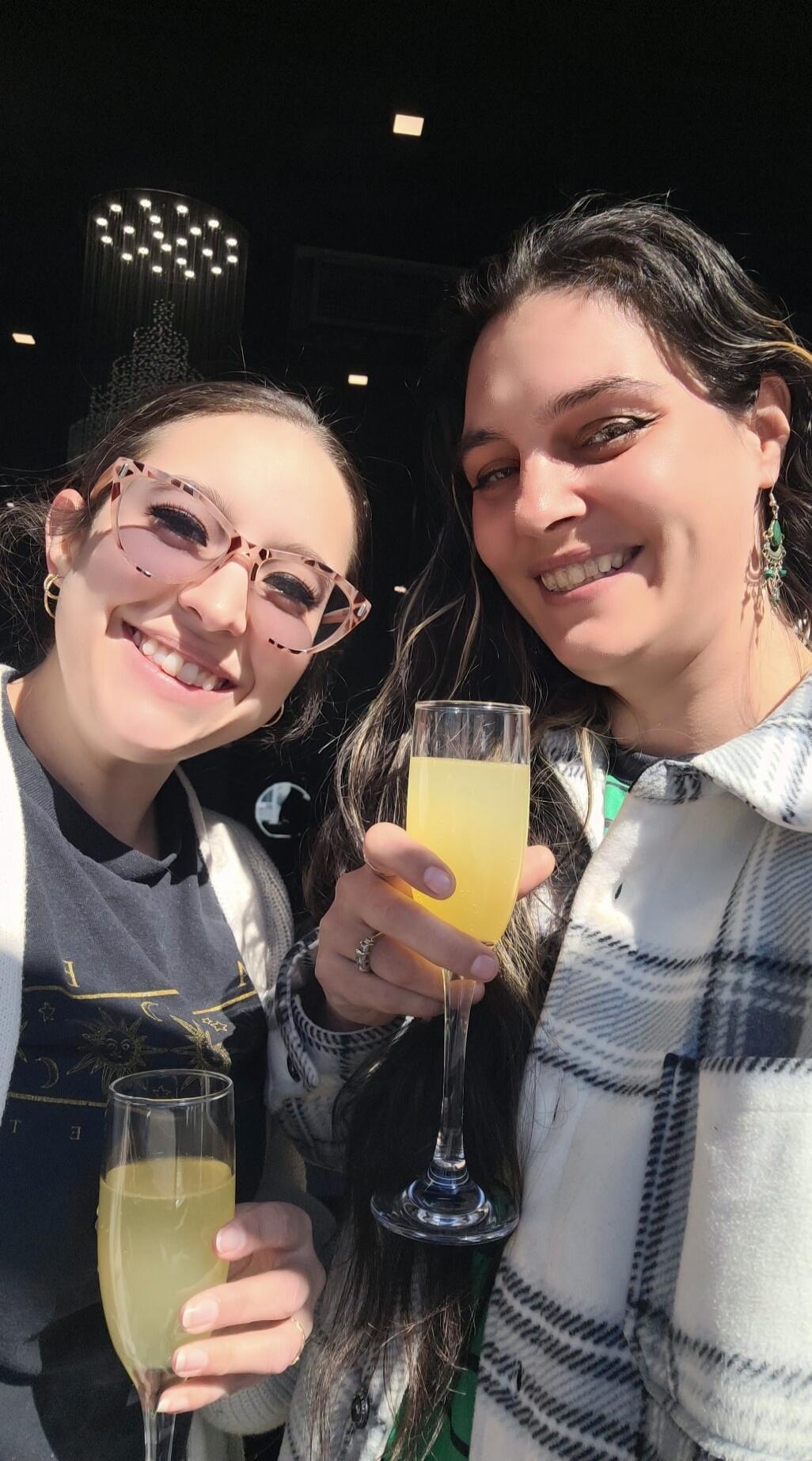 Two smiling women holding glasses of sparkling drinks, possibly mimosas, in a bright setting.