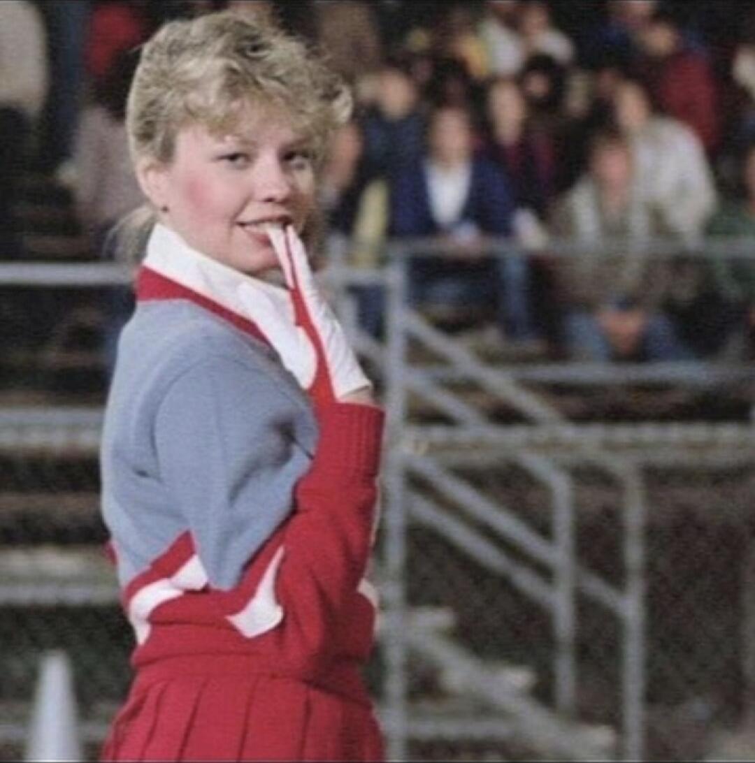 A young person in a red and blue athletic outfit is touching their lips with a gloved finger, standing in front of a chain-link fence with spectators in the background.