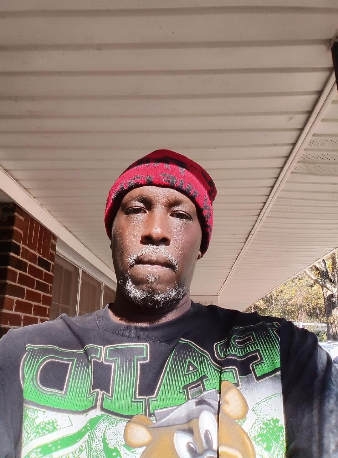 A man wearing a red and black beanie and a black t-shirt with green graphics, possibly 'GTA' and a cartoon character, takes a selfie outdoors. He has a grey beard and mustache.
