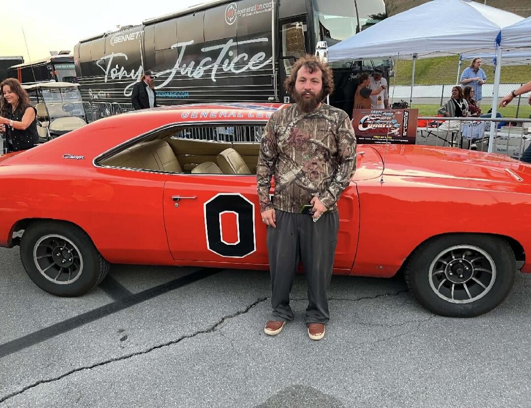 A man with a beard and a camouflage shirt stands next to an orange classic Dodge Charger, famously known as 'General Lee', with the number '01' (partially visible as '0') on its side. In the background, a black tour bus with 'Tony Justice' and 'TONYJUSTICEMUSIC.COM' written on it is visible, along with other people and white tents at what appears t