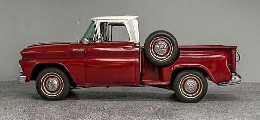 A classic red pickup truck with a white roof is parked against a plain grey background. The truck features a spare tire mounted on the side of the bed. It has chrome accents on the bumpers and hubcaps, and white-wall tires.