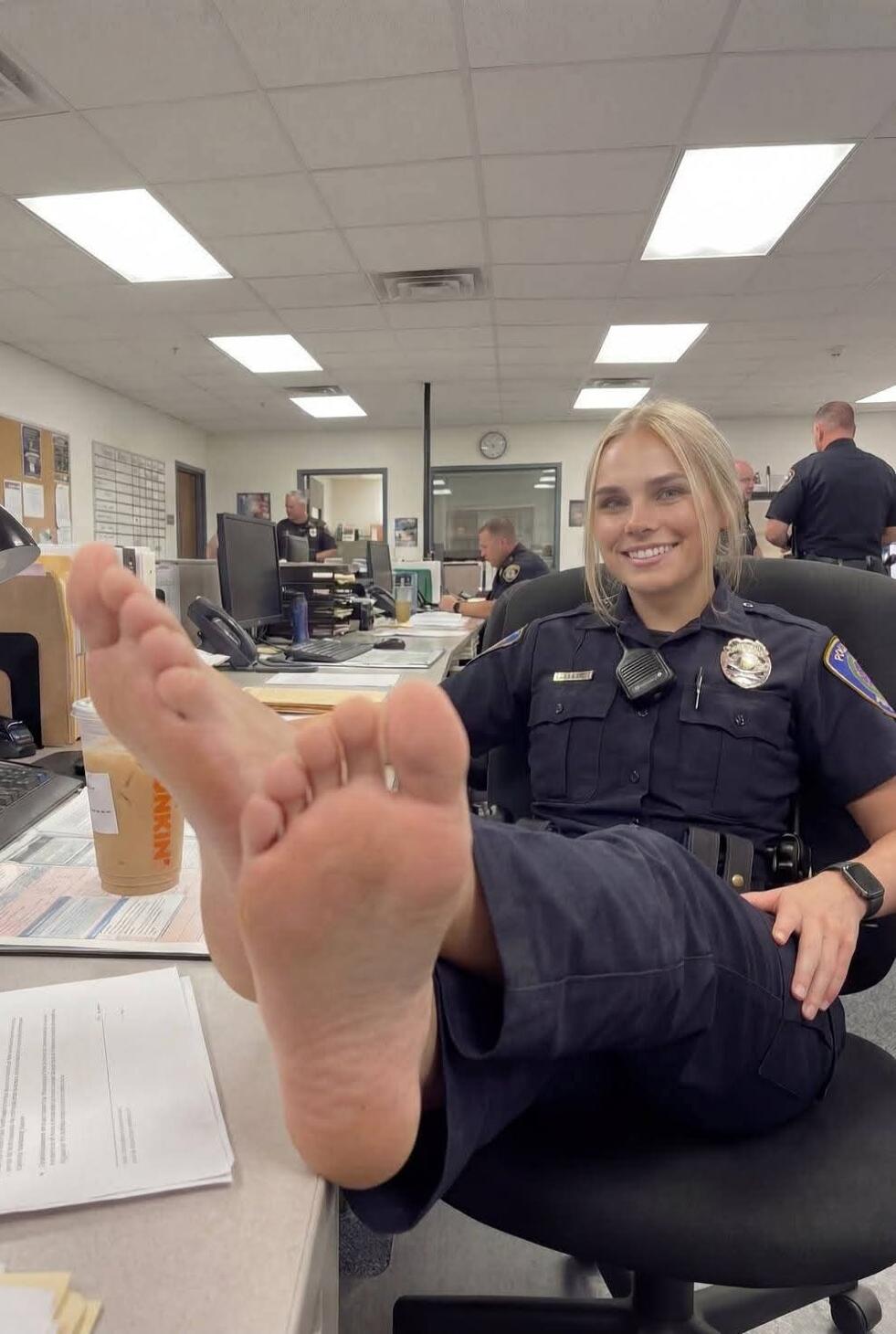 A female police officer sits at a desk with her feet up on the desk, smiling.