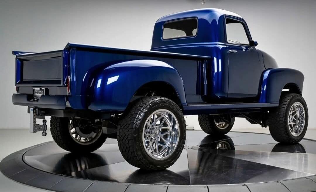 A custom classic blue pickup truck is displayed on a rotating platform, showcasing its glossy paint, large chrome wheels, and lifted suspension.