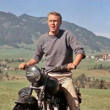 Steve McQueen riding a motorcycle in a mountainous landscape. The license plate reads WH-13371.