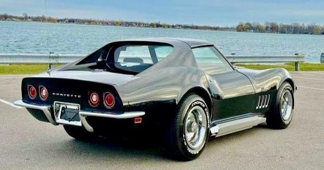 A black classic Chevrolet Corvette photographed from the rear on a lakeside road.