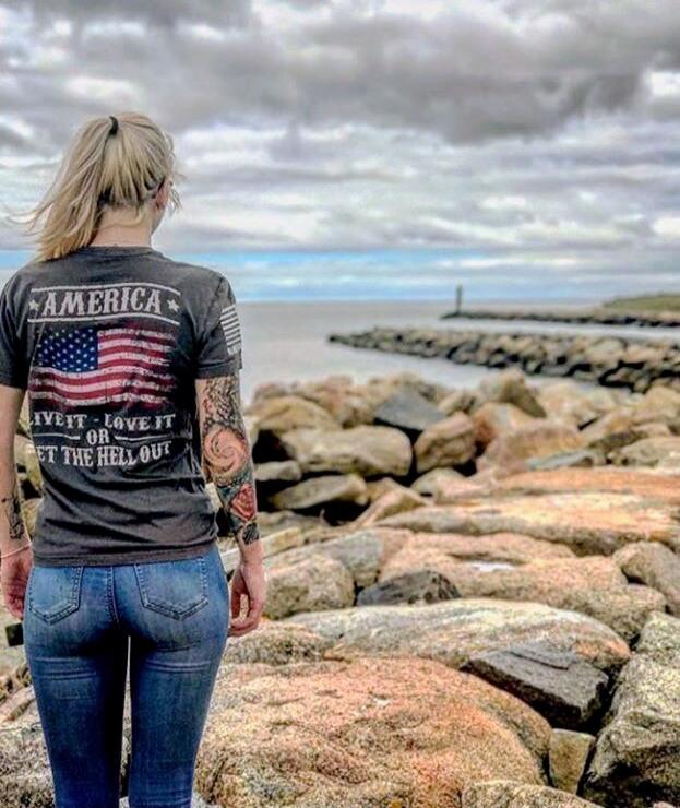 The image shows a person standing on rocky shoreline facing away from the camera. They have blonde hair in a ponytail, tattoos on their arms, and are wearing a dark t-shirt with an American flag and the words 'AMERICA' and 'LIVE IT - LOVE IT OR LEAVE IT' printed on the back, along with denim jeans.