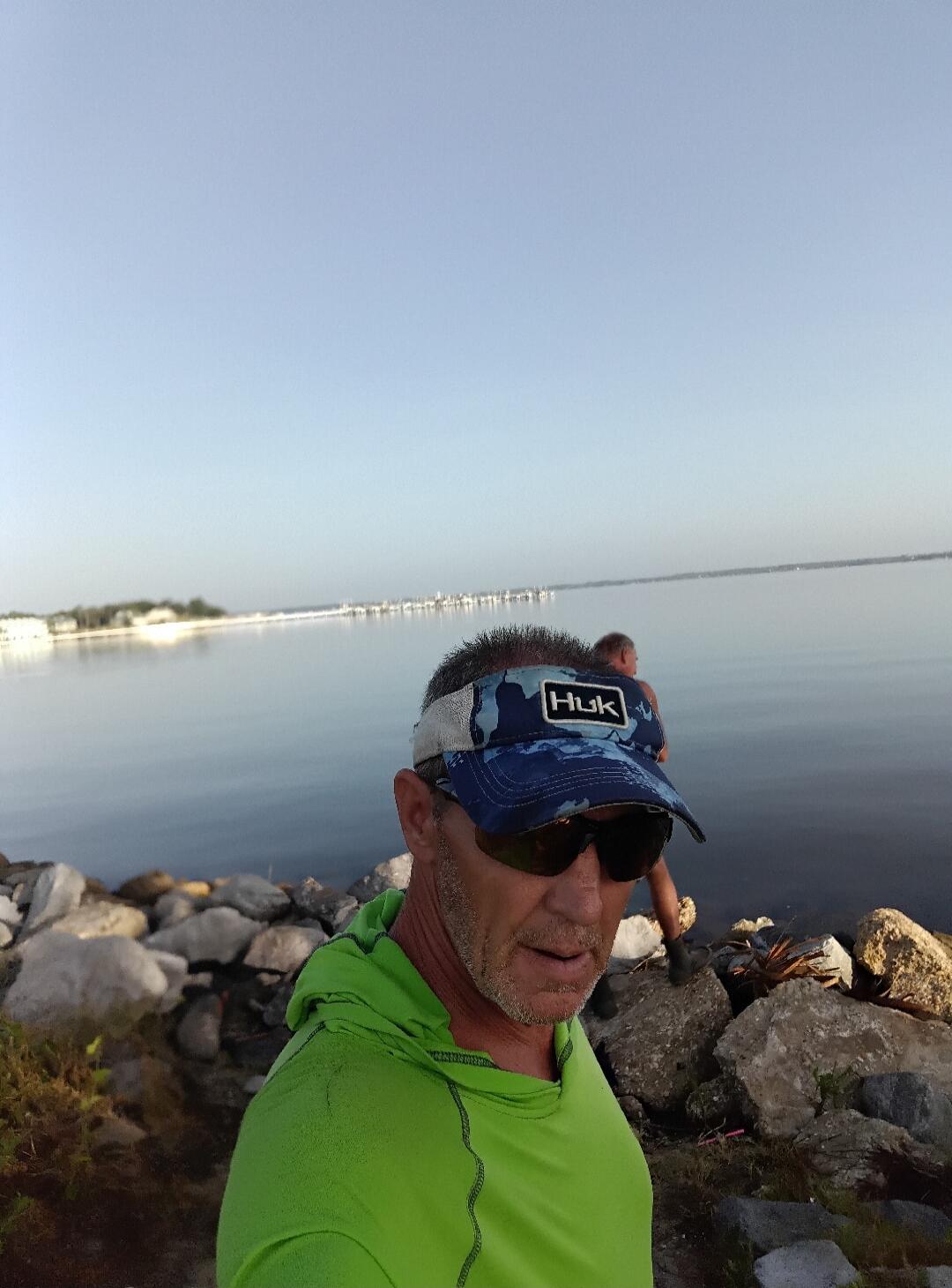 A man wearing a green long-sleeve shirt, sunglasses, and a blue camouflage visor with 'HUK' written on it, standing by a body of water with rocks in the foreground and a distant shoreline.