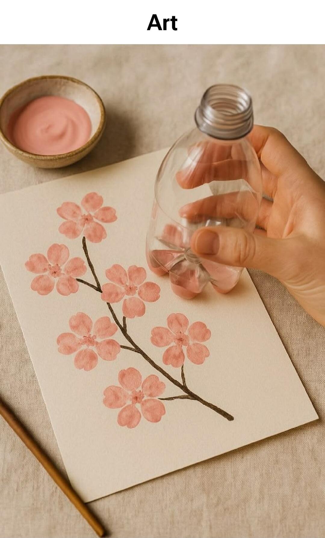 Art. A hand uses the bottom of a plastic bottle to stamp pink cherry blossom-like flowers on paper. A bowl of pink paint is nearby.