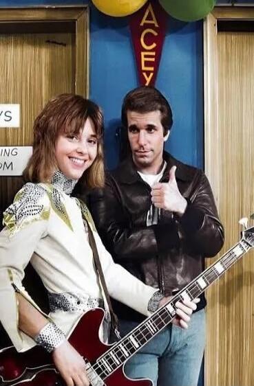 YS, ING OM, ACEY. A smiling woman in a white and gold outfit plays a red bass guitar, while a man in a leather jacket gives a thumbs-up. The scene has a retro aesthetic, reminiscent of a TV show set.