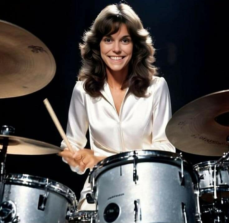 A smiling woman playing a drum set, holding drumsticks.