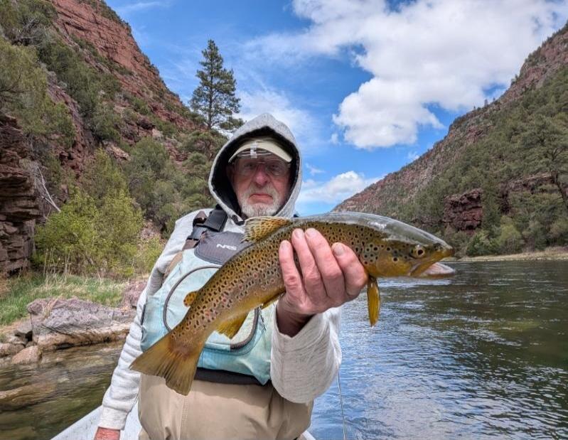 A man in a hooded sweatshirt and fishing vest holds a large brown trout he caught. He is on a river with mountains and trees in the background under a blue sky.