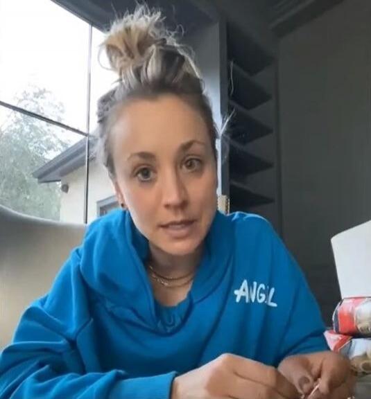 A woman with her hair in a messy bun, wearing a blue hoodie with 'ANGL' written on it, is looking at the camera and appears to be speaking. She is wearing a necklace. The background shows a window and shelves.
