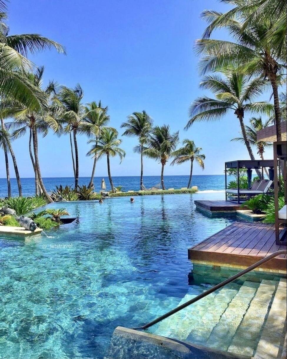 A luxurious infinity pool overlooking the ocean, surrounded by palm trees under a clear blue sky. The water is crystal clear, and there are lounge areas on a wooden deck. A watermark reads '@_jetstravel'.
