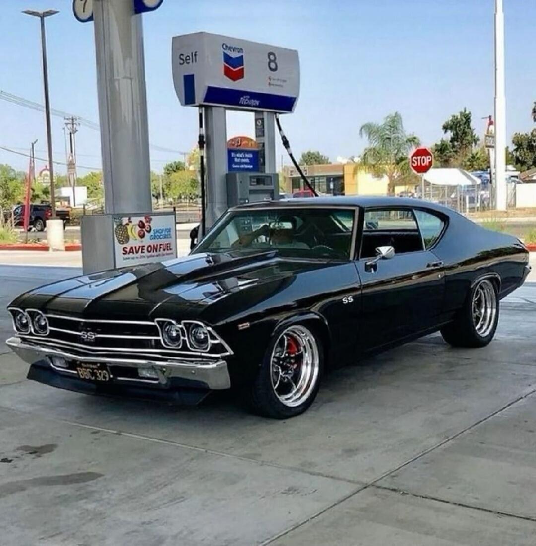 Self 8 on the gas pump. SAVE ON FUEL sign. STOP sign in the background. A black vintage muscle car parked at a gas station.