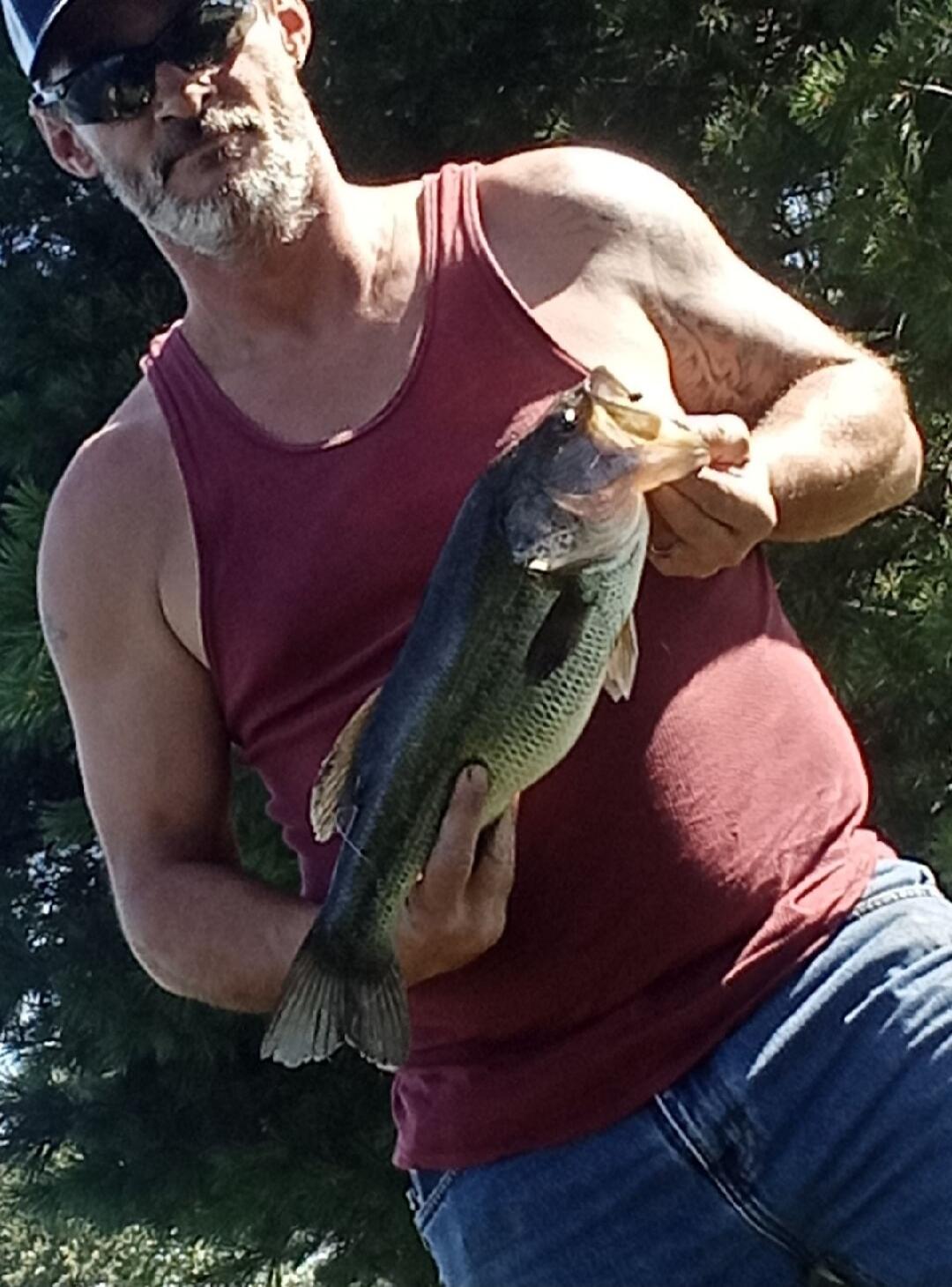 A man wearing a red sleeveless shirt holds a fish outdoors, likely after fishing.