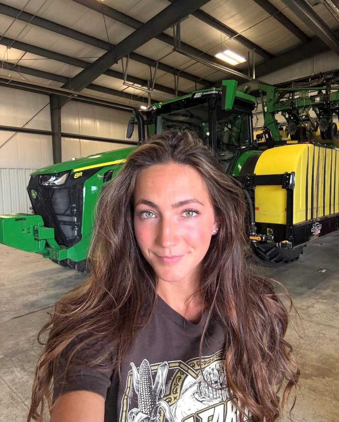 A young woman takes a selfie in front of a large green John Deere 8R tractor with yellow attachments inside a garage. Her t-shirt has a corn graphic.
