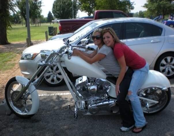 Two women posing on a white custom motorcycle in a parking lot. One woman is sitting on the tank, and the other is behind her on the seat, leaning against her. Both are smiling.