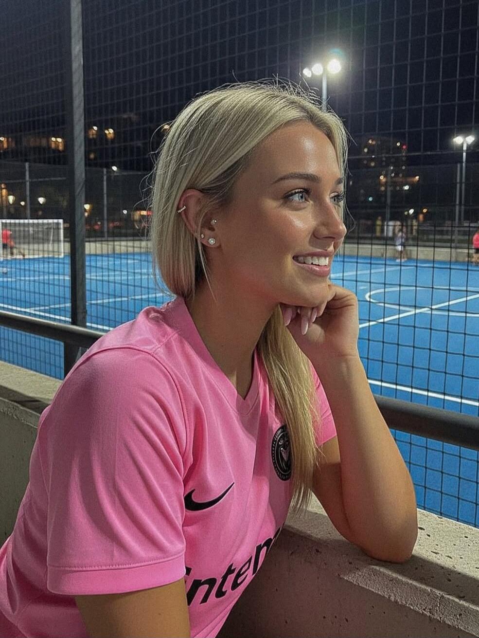 A smiling young woman with blonde hair wearing a pink sports jersey sits by a fence at a lit blue sports court at night.