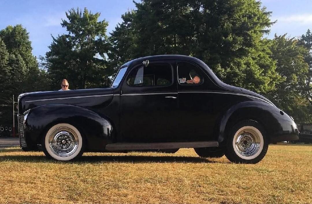 A black vintage car parked on a grassy field with people inside and around it.