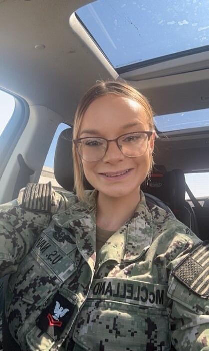 A young woman in a U.S. Navy uniform, wearing glasses and smiling, is sitting in a car. Her name 'MCCLELLAND' is visible on her chest, and 'U.S. NAVY' and rank insignia are on her sleeve. An American flag patch is also visible.
