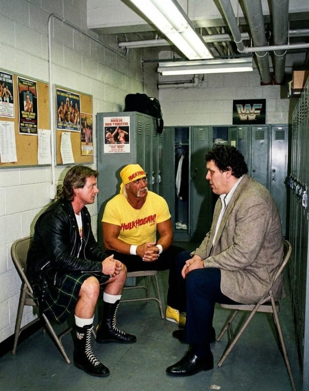 Hulk Hogan wearing a yellow shirt with 'Hulkamania' sitting in a backstage locker room with two other men (one in a leather jacket and another in a blazer). Posters on the wall and a WWF logo visible in the background.