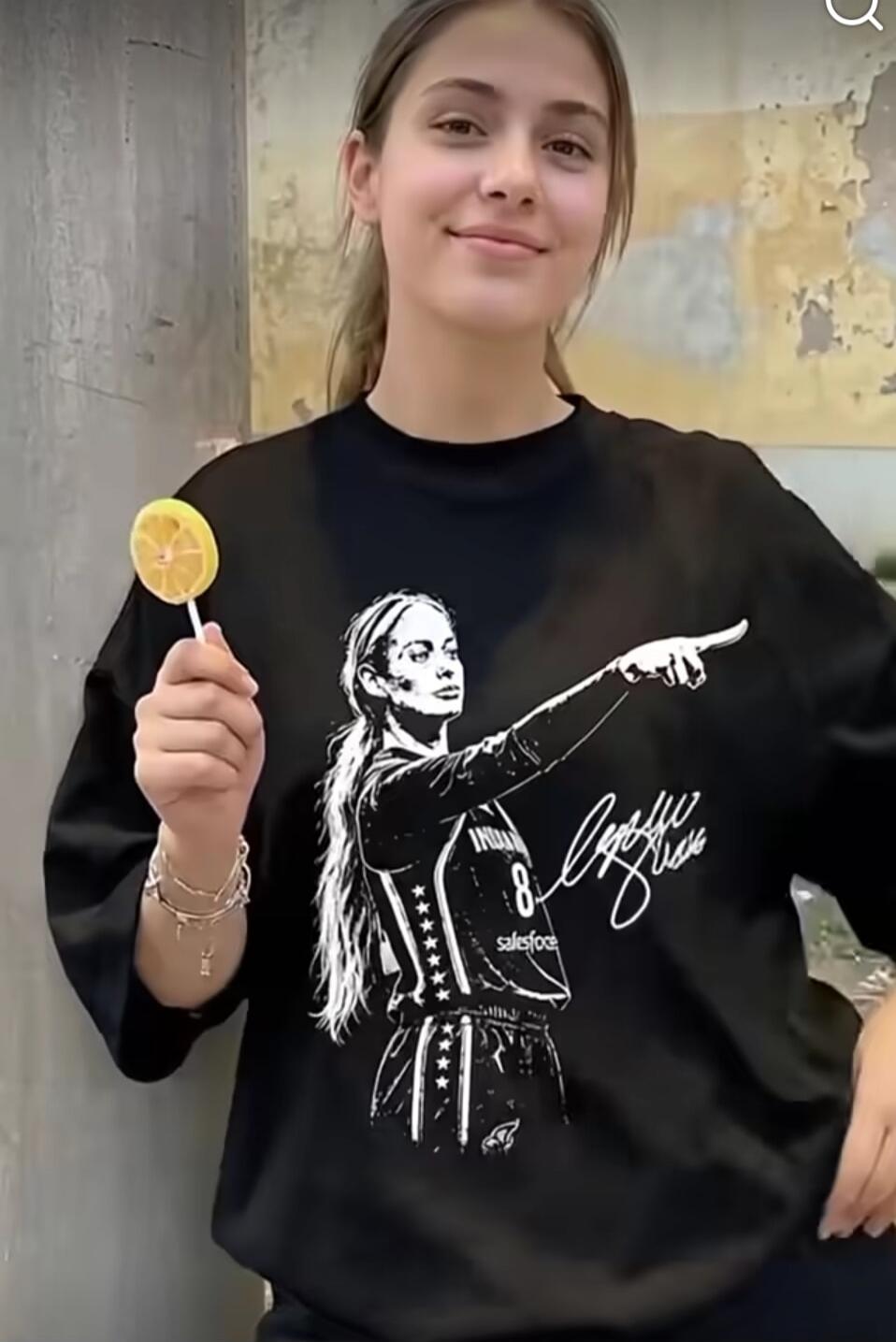 A young woman is smiling and holding a lemon slice lollipop. She is wearing a black t-shirt with a graphic print of a female basketball player, number 8, and the word 'salesforce' along with a signature.
