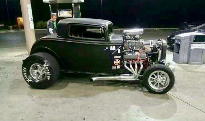 A black vintage hot rod car at a gas station with an exposed engine.
