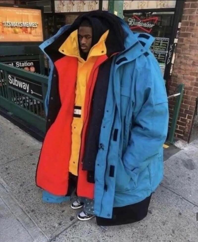 Subway sign, Budweiser sign, and a man standing on a sidewalk wearing multiple oversized jackets in bright red, yellow, and blue.