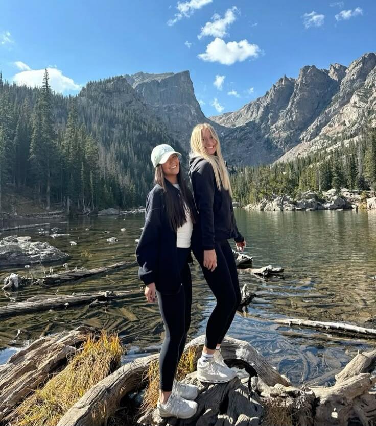 Two women standing on rocks by a clear mountain lake with forest and mountains in the background.