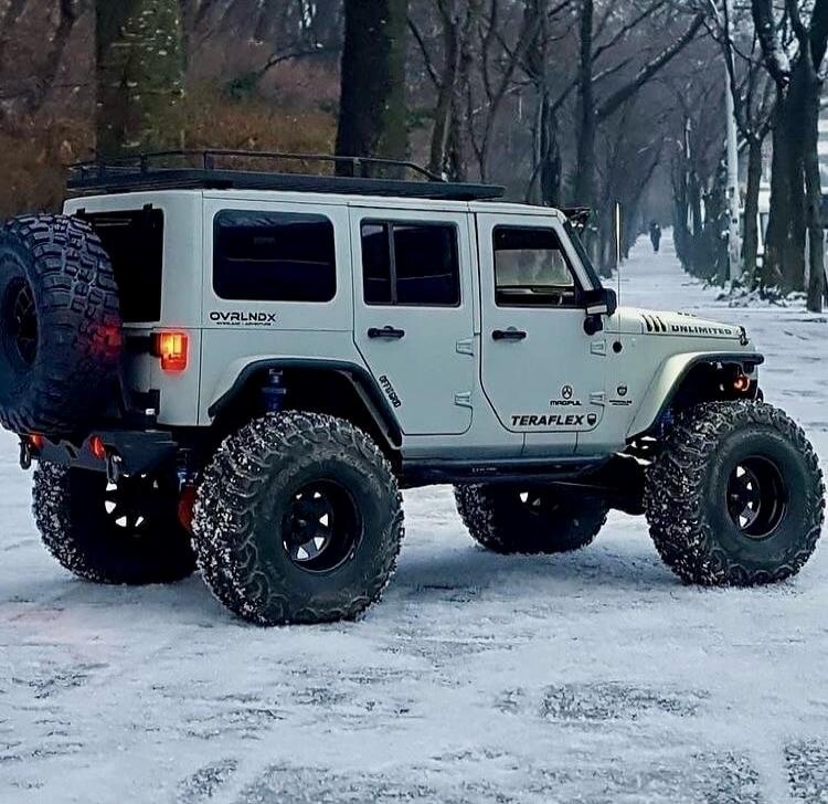 OVRLNDX OFFROAD TERAFLEX UNLIMITED MAGPUL. A modified white Jeep Wrangler with large off-road tires is parked on a snow-covered path lined with trees.