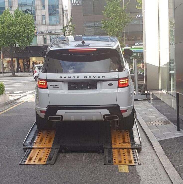 RANGE ROVER SPORT. A white Range Rover Sport SUV is seen from the rear, parked on a car lift or ramp system on an urban street.