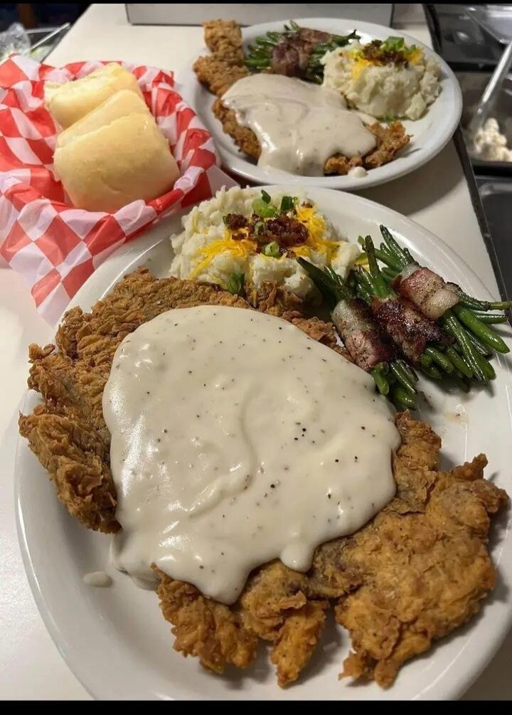 Two plates of classic American comfort food, featuring chicken fried steak covered in white gravy, mashed potatoes with cheese and bacon bits, and green beans wrapped in bacon. A basket of dinner rolls is also visible.