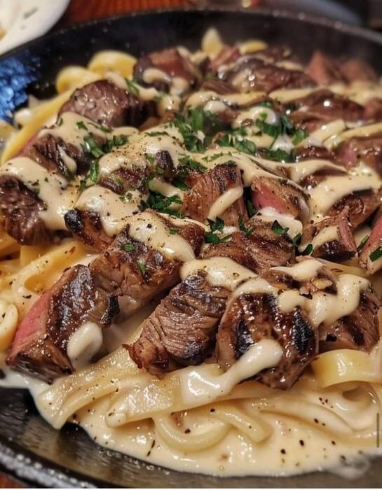 A pan of pasta topped with sliced grilled steak, creamy sauce drizzle, and herbs. The dish appears to be fettuccine or tagliatelle with cubes of beef and a light herb garnish.