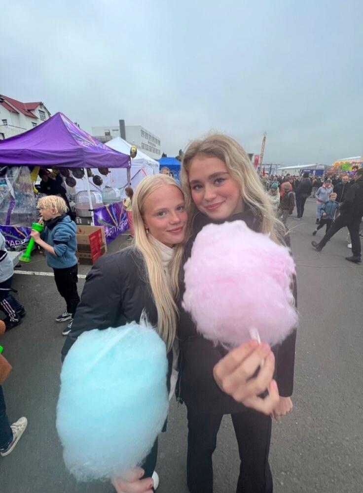 Two young women are smiling and posing for a photo at an outdoor event, each holding a large cloud of cotton candy. One woman has pink cotton candy, and the other has blue. In the background, there are tents, other people, and a general fair or festival atmosphere.