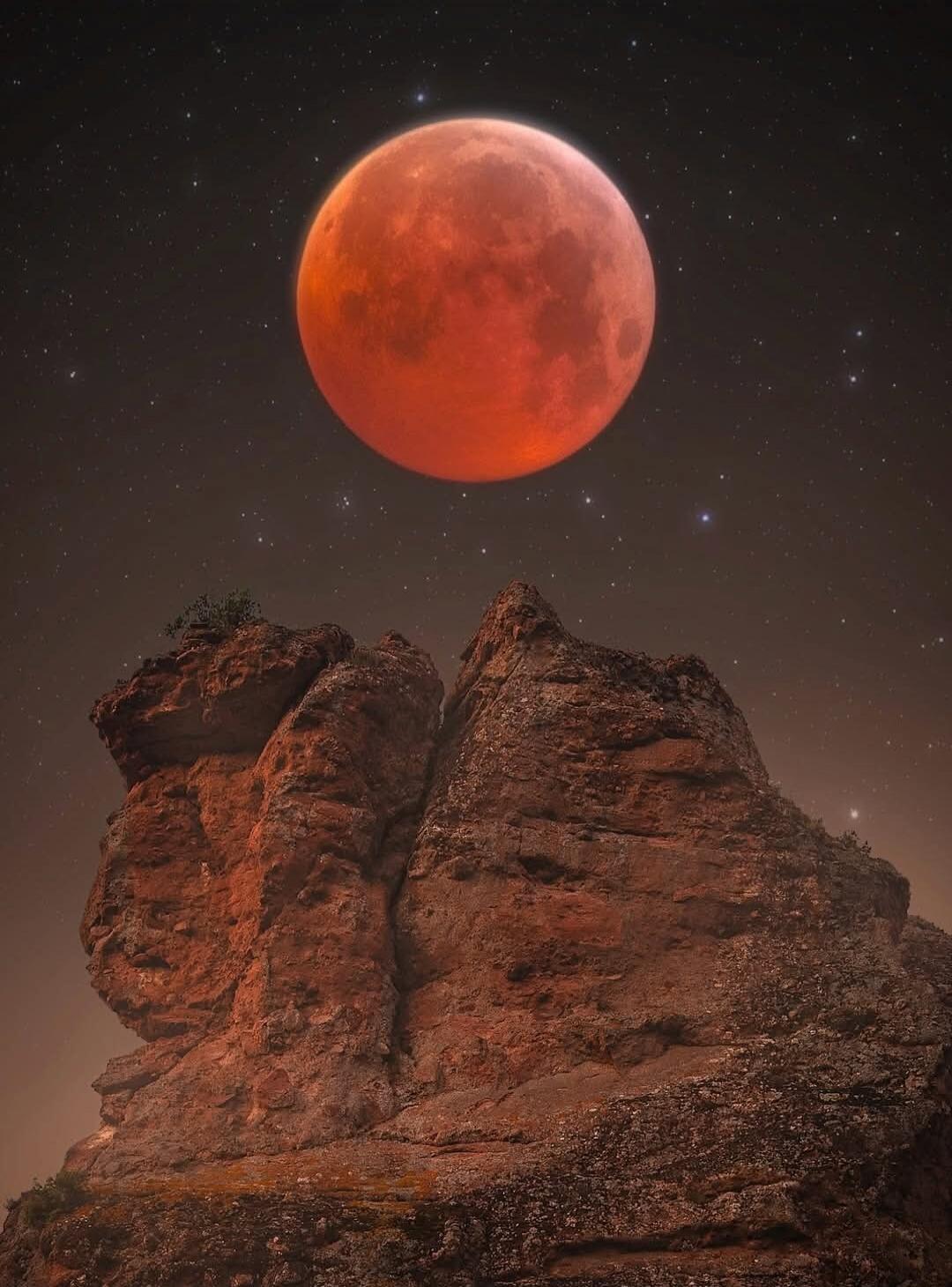 A red lunar eclipse visible over rugged rock formations.