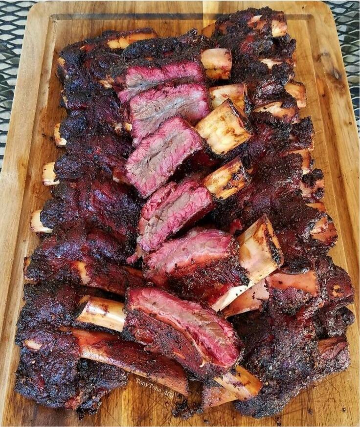 Stack of smoked barbecue ribs on a wooden cutting board, sliced to show juicy pink interior with a dark, bark-like crust.