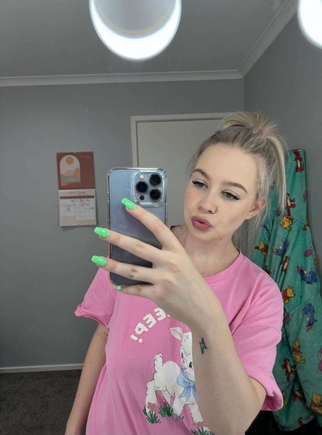 A young woman with blonde hair in a ponytail takes a mirror selfie. She is wearing a pink t-shirt with a lamb graphic and has bright green nails. She holds an iPhone to take the picture. In the background, there's a grey wall, a calendar, and a blanket with cartoon characters.