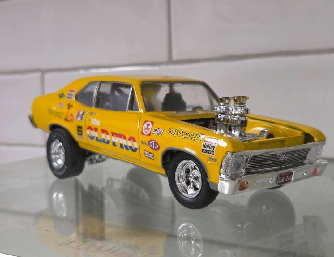 A yellow model car, possibly a Chevrolet Nova, customized for drag racing. It features an exposed engine with prominent chrome carburetors or blowers. The side of the car has various decals including 'the OLD PRO', 'GROUCHY', 'STP', 'Champion', 'V', and 'LEE'. The front bumper has a license plate that reads 'CJO-214'. The car is also marked 'Holley