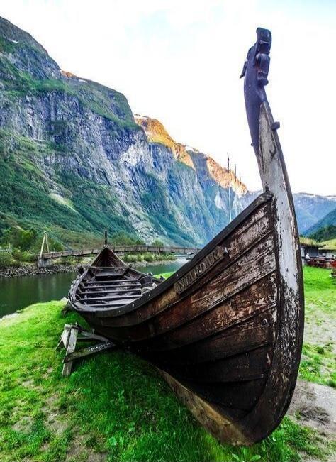 Two old wooden boats, resembling Viking longships, are resting on a grassy bank next to a calm body of water. In the background, majestic green mountains rise under a bright sky. A wooden bridge spans the water. The name 'MARDAR' is visible on the side of one boat.