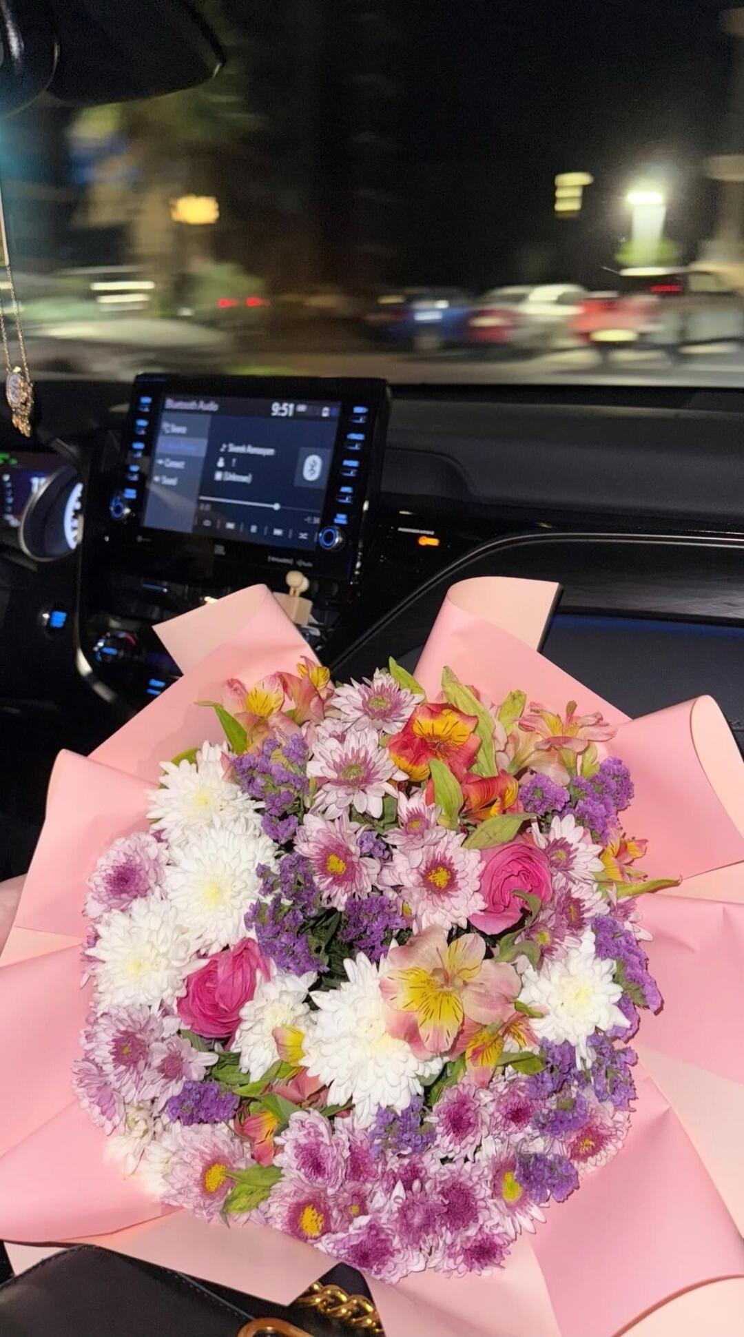 A vibrant bouquet of mixed flowers is placed inside a car at night. The car's infotainment system shows '9:51', 'Bluetooth Audio', and other menu options.