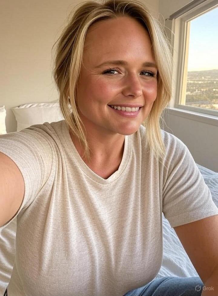 Smiling woman taking a selfie in a light room by the window. She has blonde hair and is wearing a beige t-shirt.