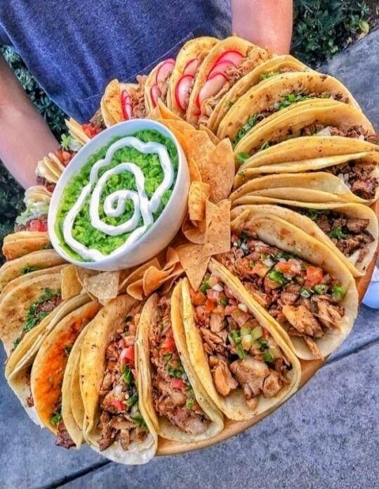 A round platter of many tacos filled with meat and vegetables, arranged in a circular pattern with a bowl of green sauce in the center.