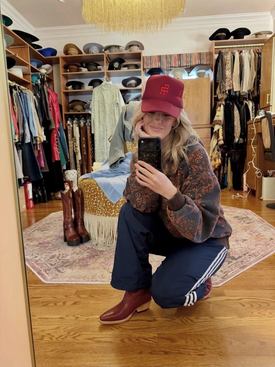 photo of a person kneeling in a walk-in closet taking a mirror selfie wearing a red cap, a patterned sweater, navy pants with white stripes, and red ankle boots.
